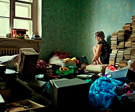 Une jeune femme en fond de plan assise sur un tas d'accumulations dans son appartement lui-même remplit d'accumulations